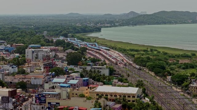 Aerial drone view showing multiple trains positioned on parallel railway tracks beside a broad waterbody. Buildings trees and distant hills surround the railway yard in Chennai India