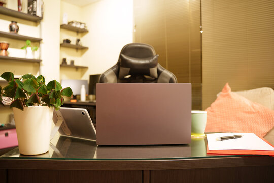 Home office desk with closed laptop, potted plant, notebook, mug, pen, cushioned chair, and warm lamp light beside textured wall paneling, creating calm productive workspace