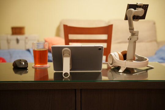 Tablet on stand beside smartphone and compact robotic arm on living room table, with tea glass and mouse behind, warm lamp light reflecting across glossy dark cabinet surface