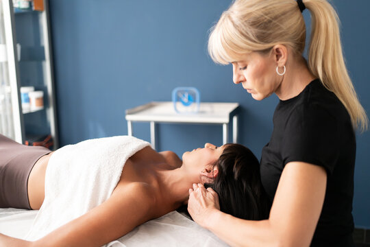Female therapist performing neck and shoulder massage on relaxed adult woman in treatment room with soft natural light
