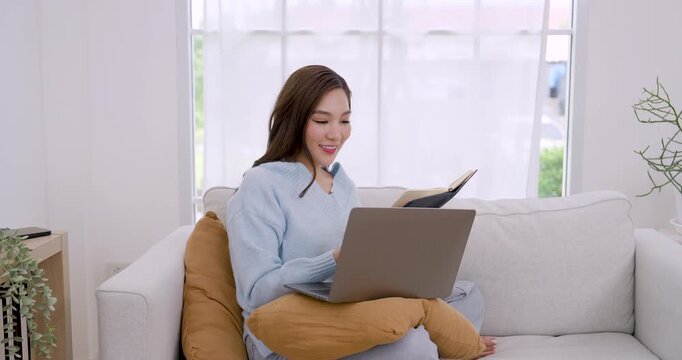 Young Asian woman works from home on sofa with laptop and headphone, listening music while remote working, smiling, moving hand to rhythm during stress relief break in bright cozy minimalist apartment