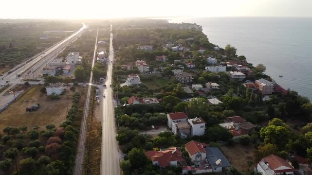 Drone aerial view over Alissos near Patras, Greece, revealing a coastal road, the blue Ionian Sea and the town beneath a dramatic sunset sky, creating a peaceful Mediterranean atmosphere
