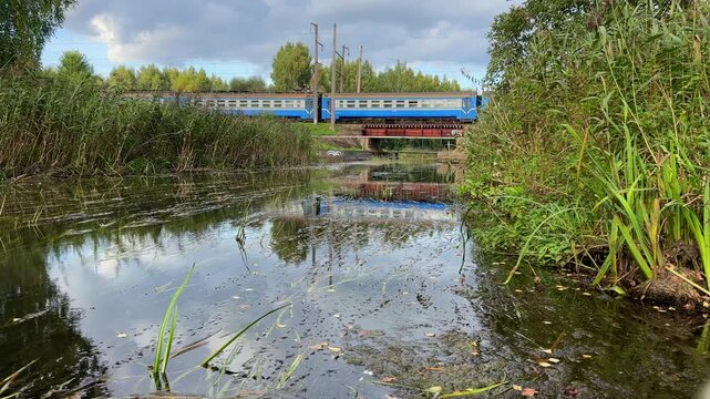 Electric train on railway bridge crossing a small, overgrown canal. Blue passenger train moves across bridge against backdrop of summer or early-autumn landscape. Travel on passenger train. 