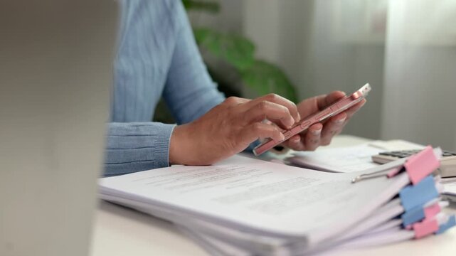 Dedicated female accountant reviewing printed paper report while typing on modern smartphone balancing corporate budget and verifying accounts in comfortable indoor office room