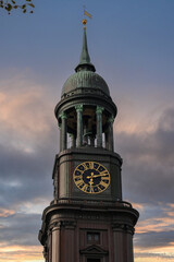 A striking architectural vertical shot of a historic European church clock tower featuring a classical gold Roman numeral dial, decorative dark green copper cupola dome, open colonnade balcony © Victor