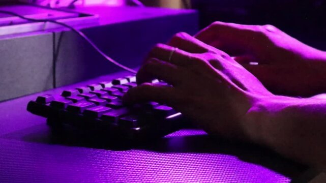 A person types on a computer keyboard at a desk illuminated by purple ambient light to work late at night.