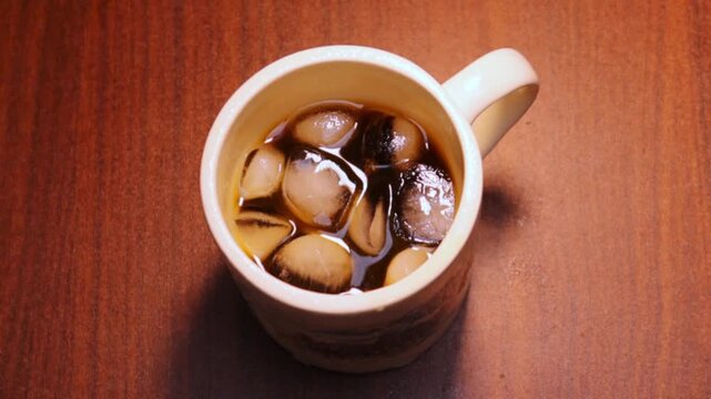 Ice cubes float inside a ceramic mug filled with iced coffee placed on a wooden table.