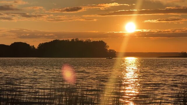 Boat moves slowly along river on sunset scene in evening. Sunset over lake, casting light on its surface. Fishing boat passing by against backdrop of wooded island in distance. 