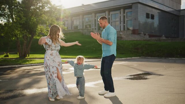 Family of three dances joyfully in a sunlit driveway. The mother twirls, laughing with delight. The father claps, smiling broadly. Their toddler runs between them, arms outstretched