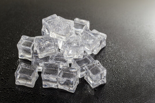 A pile of clear, transparent ice cubes rests on a dark, smooth surface, showing their frozen crystalline structure.
