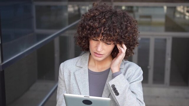 A young woman sitting outdoors, casually utilizing a tablet in a modern urban setting