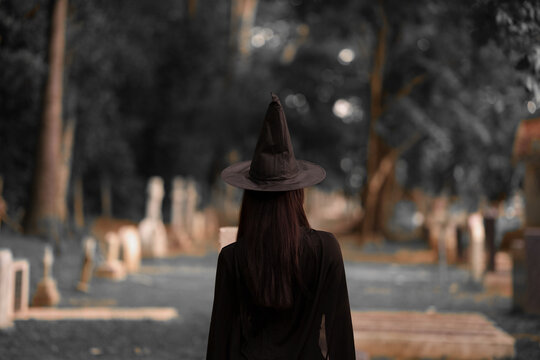 Back view of a mysterious witch in a black pointed hat and flowing cape, standing in a dark forest cemetery with an eerie Halloween atmosphere.