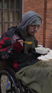 Vertical video of homeless Caucasian young adult man with disability eating instant noodles in wheelchair outdoors. Clip suitable for charity campaigns and social issue awareness projects