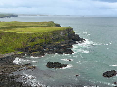 Grassy sea cliffs rise above dark volcanic rocks and Atlantic waves along the rugged Causeway Coast near Giant's Causeway in Northern Ireland.