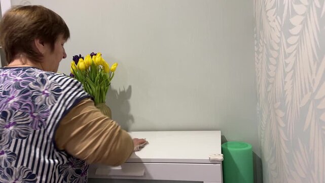 Side view of mature woman arranging golden vase with colorful tulips on white chest of drawers