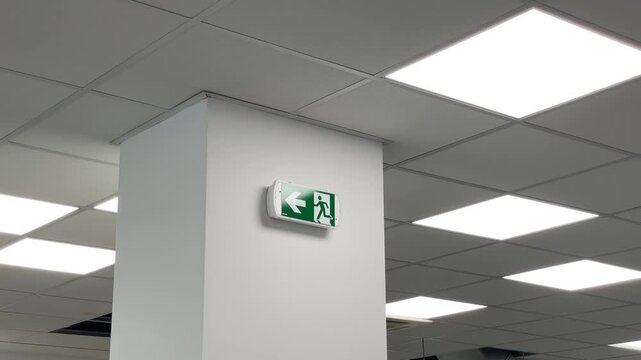 Interior view of suspended office ceiling grid with recessed light panels, white pillar and glowing exit sign, showing clean modern commercial space