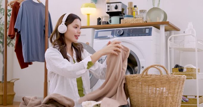 Young Asian woman folds beige and white clothing beside washing machine, smiling softly with headphone on, listening music while finishing laundry task in clean minimalist apartment interior