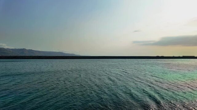 Serene coastal landscape featuring a long stone breakwater protecting the calm blue sea harbor during a beautiful sunset with distant mountain silhouettes under a clear vast sky at the seaside.