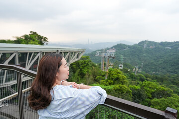 Obraz premium A side-profile shot of a young Asian woman leaning against a railing at an elevated observation platform, taking in the panoramic landscape of green hills and the Maokong Gondola cable car line in Tai