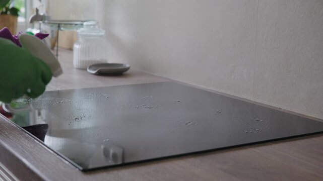 Person in rubber gloves spraying cleaning detergent onto a ceramic induction hob and wiping it clean with a microfiber cloth in a kitchen.