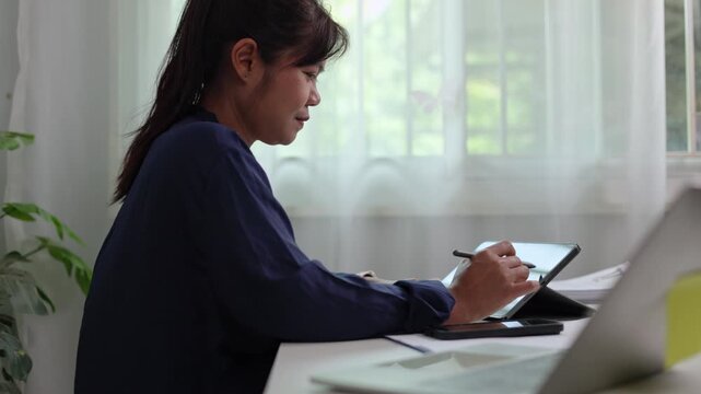 Focused woman drafting strategy document on slim tablet device with pen stylus while seated near window inside peaceful corporate office room during daytime working hours