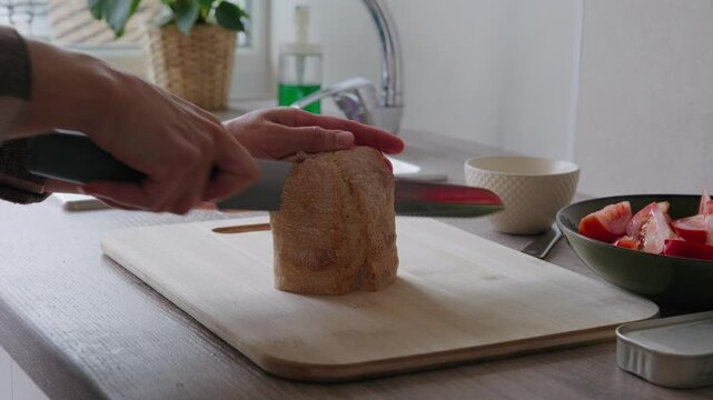 Close-up of a person slicing bread on a cutting board and spreading cream cheese on it in a kitchen.