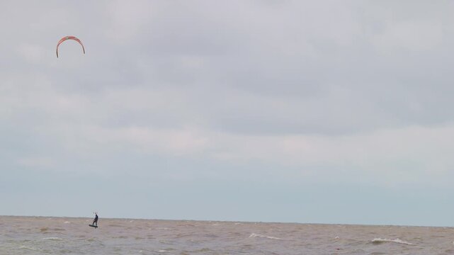 Action wide shot of a lone kitesurfer with kite gliding across rough ocean waters on an overcast day. Extreme water sports and active lifestyle concept. Flying over water in slow motion..