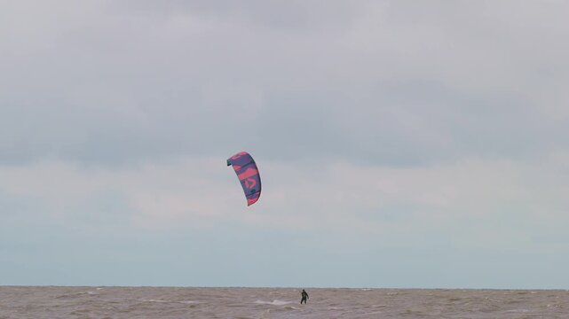 Action wide shot of a lone kitesurfer with kite gliding across rough ocean waters on an overcast day. Extreme water sports and active lifestyle concept. Flying over water in slow motion..