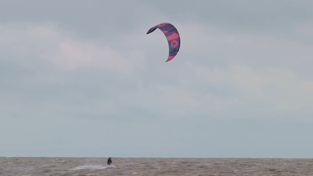 Action wide shot of a lone kitesurfer with kite gliding across rough ocean waters on an overcast day. Extreme water sports and active lifestyle concept. Flying over water in slow motion..