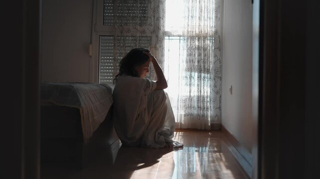 Depressed young woman sitting on the floor beside her bed wrapped in a bedsheet near a window, experiencing sadness, loneliness and emotional distress at home.