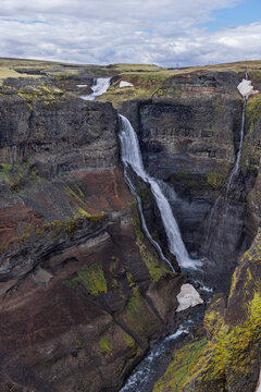Granni waterfall on the Fossa River in Thjorsardalur, Iceland, bends down layered cliffs into a deep gorge, with snow patches, green vegetation and a winding stream below the stepped falls.