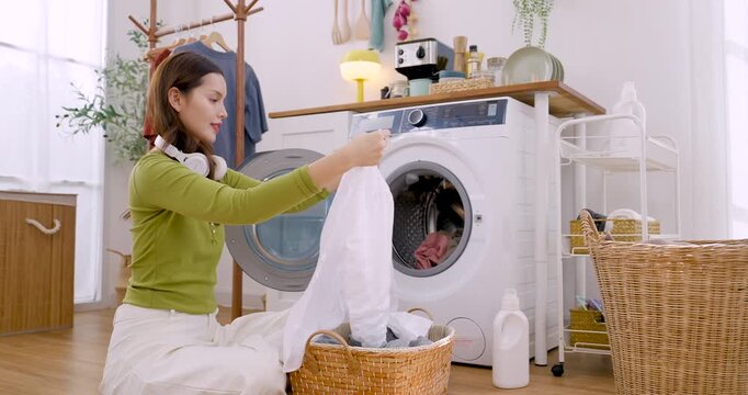 Young Asian woman kneels at washer, holds soft garment, places laundry inside drum, organizing textile care during calm housekeeping routine in cozy bright apartment with clean interior today