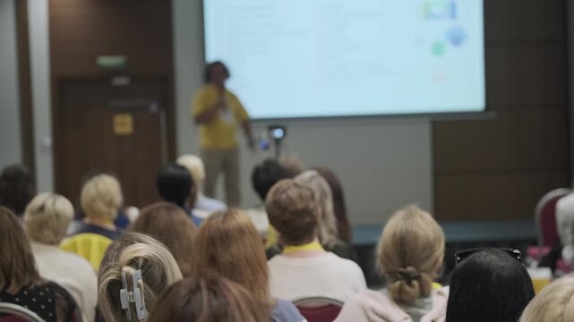 Conference audience listening to speaker during presentation, blurred presenter and projection screen, professional seminar, corporate training atmosphere.