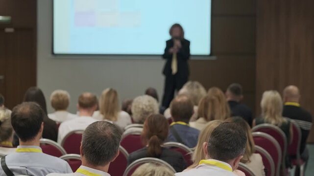 Business conference audience listens to keynote presentation in corporate seminar room, focus on attendees from rear view, professional networking and learning atmosphere.