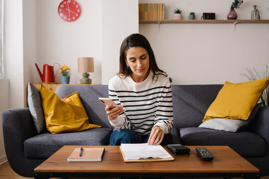 Woman using smartphone while organizing financial family documents at home