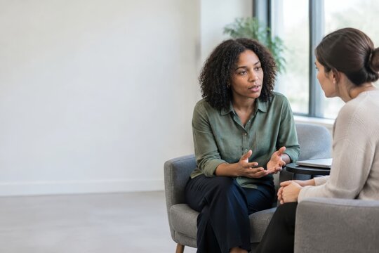In a contemporary room adorned with expansive windows that let in ample daylight, two women are deeply engaged in an animated conversation, with a leafy plant adding a touch of nature to the backgroun