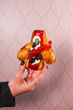 Hand holding a decorative laminated pastry topped with red berry glaze, cream, red currants and blackberries against a soft pink background.