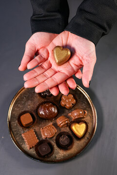 Hands holding a heart-shaped golden chocolate praline above an assortment of handmade chocolates and bonbons arranged on a decorative tray.