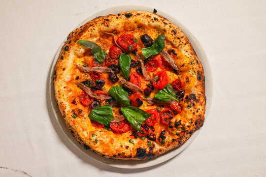 Top view of whole Neapolitan-style pizza with cherry tomatoes, anchovies, black olives, capers and fresh basil on a light background.