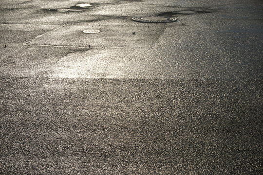 Graue nasse asphaltierte Stra&szlig;e mit Gullideckeln und Licht bei Sonne und Regen am Abend im Sommer