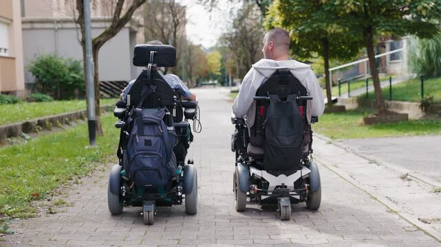 Two powered wheelchair users enjoying a conversation while moving along an accessible sidewalk in an urban environment.