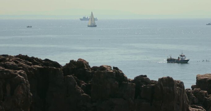 View of Portugal&rsquo;s  Atlantic coastline with dark rocks in the foreground and a variety of vessels &mdash; including a fishing boat, yacht, and ship &mdash; sailing in the brightly sunlit ocean in the background.