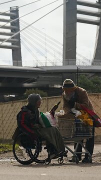 Vertical shot of senior Caucasian homeless man handing bottle to man in wheelchair with disability outdoors. Clip fits charity campaigns and social welfare documentaries