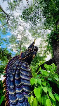 Colorful Naga serpent sculpture beside a large tree in a lush outdoor setting, with intricate scales and vivid details surrounded by green foliage and bright blue sky.