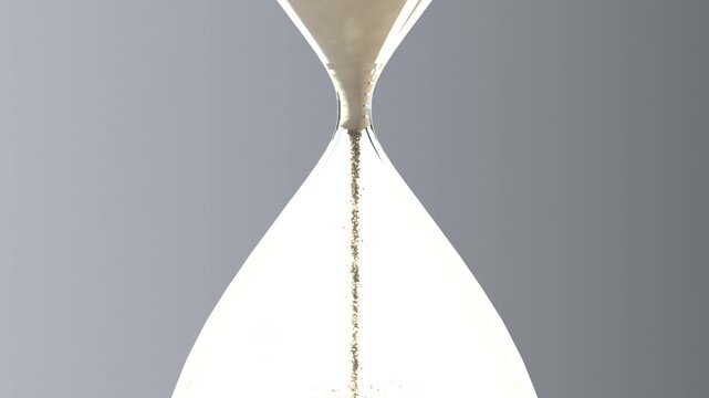 Emptying top bulb clear hourglass releasing tan sand into lower bulb forming conical pile in studio