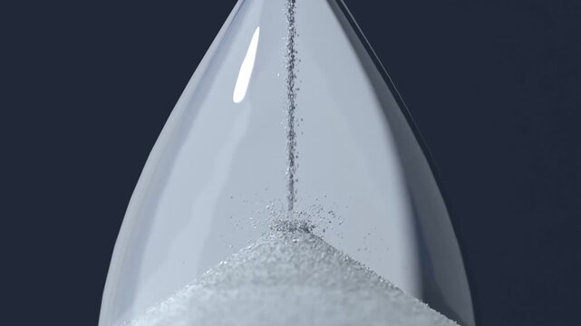 Flowing fine white sand falling by gravity through hourglass neck, building conical pile in studio