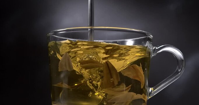 Hot green tea stirring with spoon in glass cup with steam on black background in slow motion