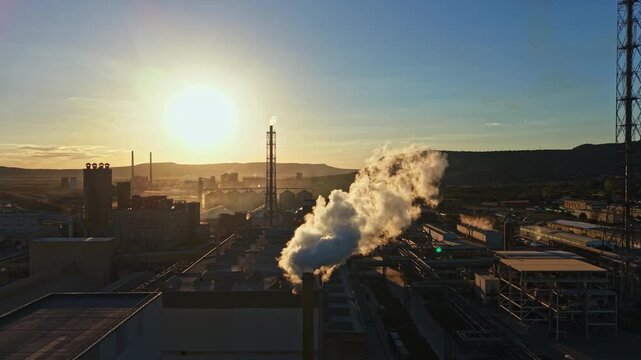 Smoke is seen coming from a tall stack at an industrial plant during sunset. The sky is bright with hues of orange and yellow. Buildings and structures are visible below.