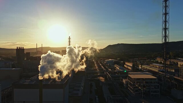 Smoke rises from factories in an industrial area as the sun rises in the background. The scene captures the start of a workday in a manufacturing zone.