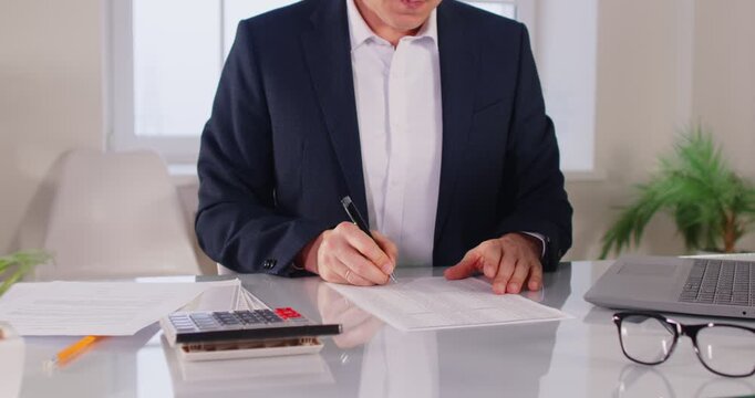 Cropped view of man in formal suit sitting at desk, working with papers and looking through financial documents. Male accountant or businessman reviewing accounting data during workday in office.